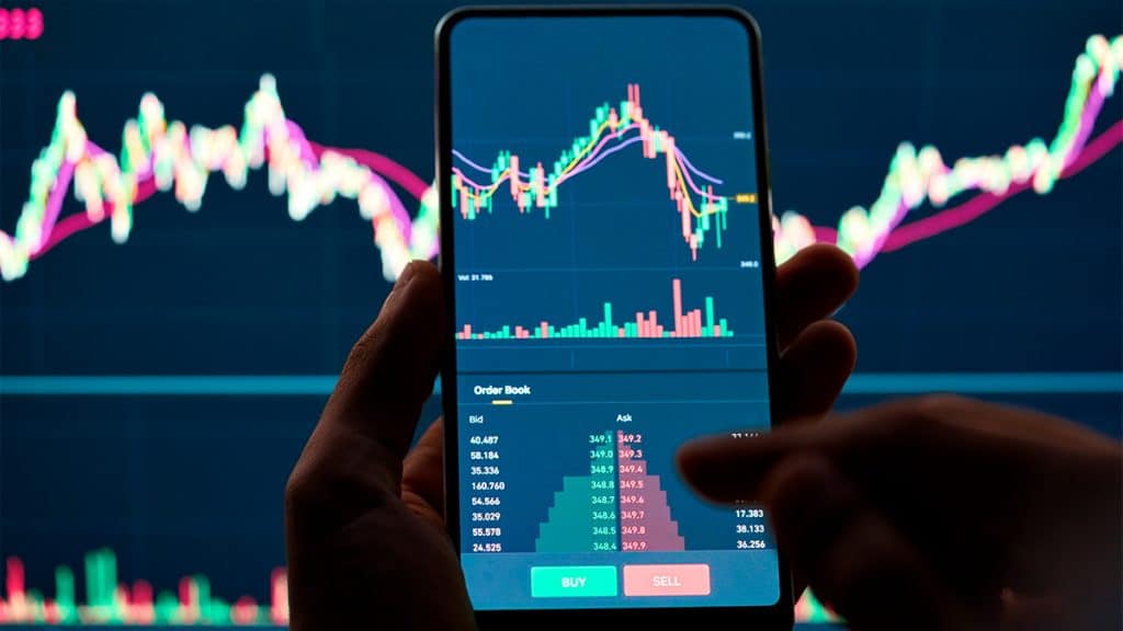 Pessoa com aplicativo de investimentos aberto em celular na frente de monitor com linhas de ações financeiras