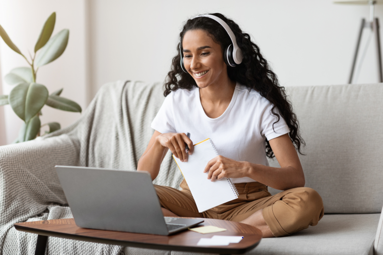Mulher jovem sentada em um sofá, segurando um bloco de notas enquanto assiste uma aula remota em um notebook. Foto: Getty Images
