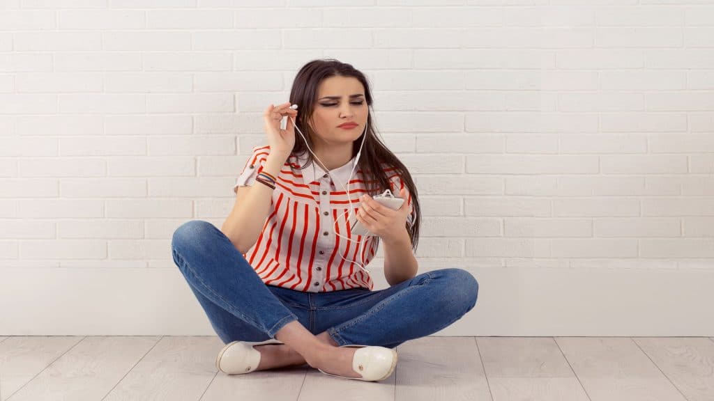 Jovem com os olhos fechados, irritada com o que ouviu nos fones, segurando o celular e ouvindo um audiobook que está muito alto, sentada de pernas cruzadas. Fundo de parede de tijolos brancos isolados.