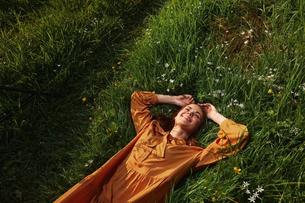 uma mulher feliz e relaxada, deitada na grama verde, usando um vestido longo laranja, com os olhos fechados e um sorriso agradável no rosto, aproveitando a harmonia com a natureza e se recuperando