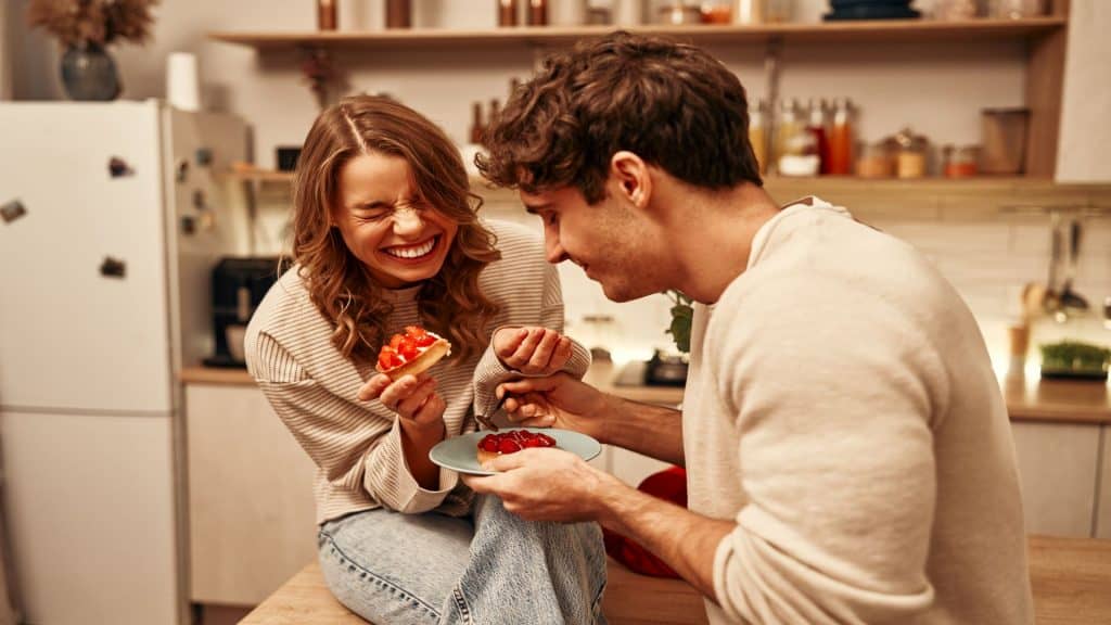 Casal jovem apaixonado comendo bolos enquanto estão sentados à mesa na cozinha, passando a noite romantica juntos.