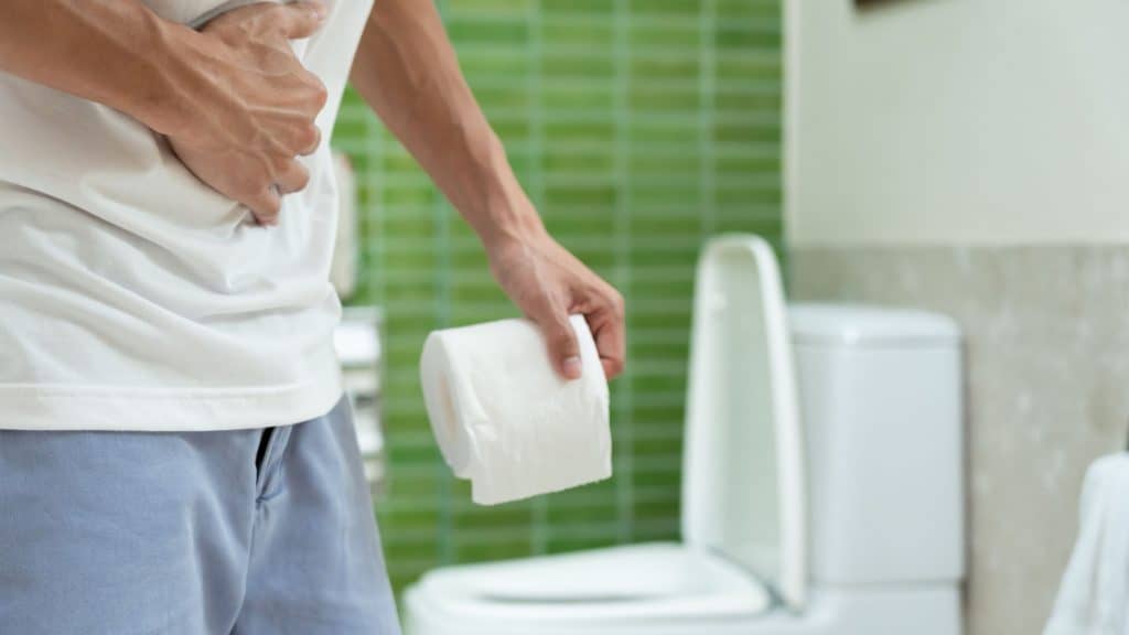 homem cor dor de barriga, segurando papel higienico, de pé, em um banheiro