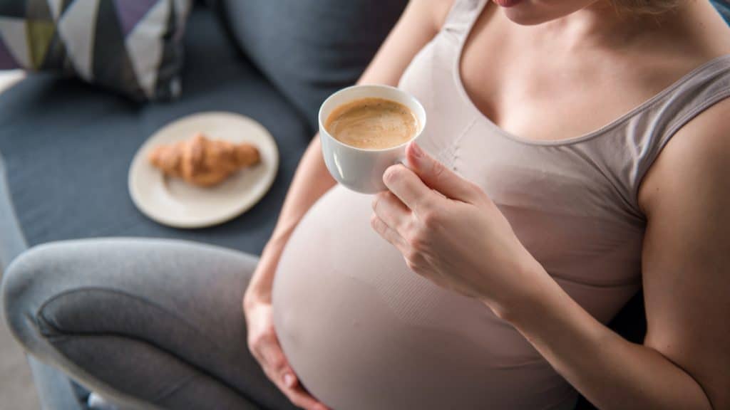 Mulher grávida tomando café, sentada no sofá e passando a mão na barriga
