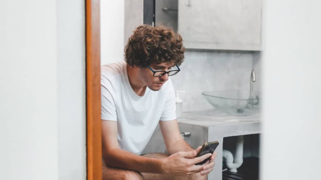 Jovem gamer caucasiano sentado na privada no banheiro jogando online no smartphone. Vício, dependência, hábito, rede social, mídia, tecnologia. Homem usando o telefone.