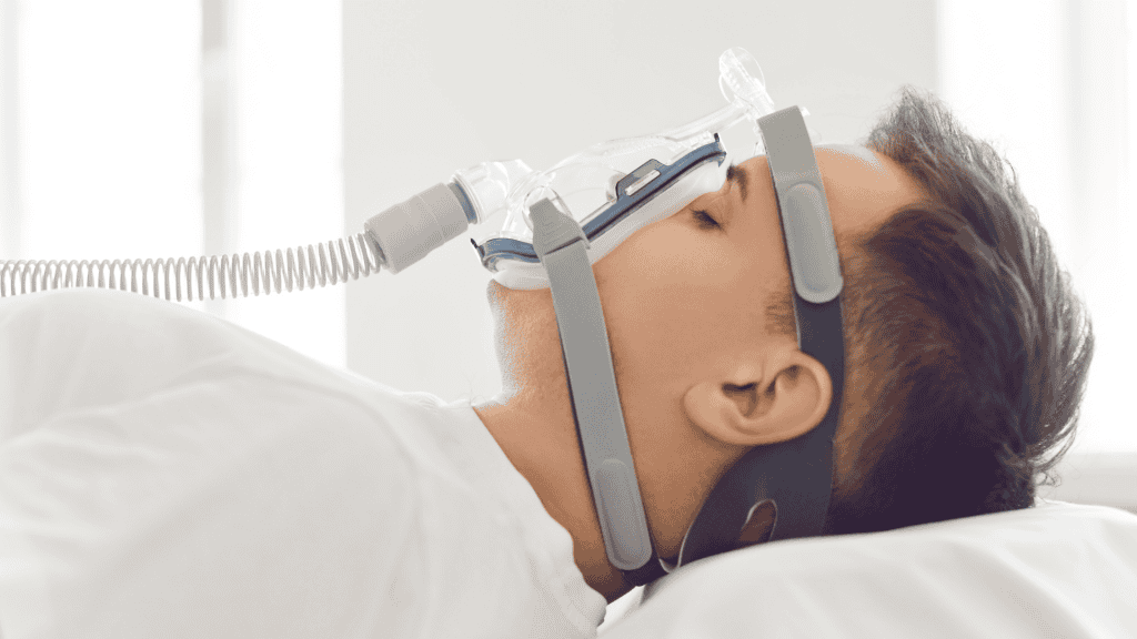 Vista lateral próxima de um paciente do sexo masculino dormindo em uma cama de hospital com máscara de oxigênio no quarto branco. Jovem se recuperando após a doença ou recebendo terapia de apneia obstrutiva do sono na enfermaria do hospital.
