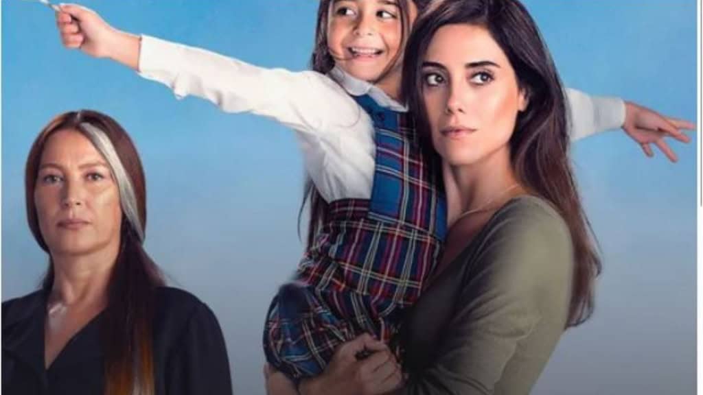 Três pessoas posam diante de um fundo azul: à esquerda, uma mulher de cabelo castanho com mecha branca veste preto e olha séria. No centro, uma menina sorridente de uniforme escolar é erguida por outra mulher à direita, que tem cabelo escuro e expressão séria, vestindo verde.