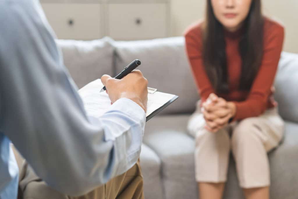 mulher sentada em sofá na frente de médico com prancheta, que faz anotações