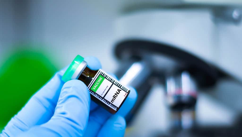 Mão com luva segura um frasco identificado como vacina de mRNA em primeiro plano, com equipamentos de laboratório desfocados ao fundo.